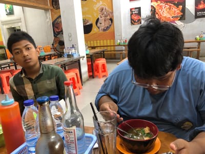 The image depicts an indoor dining scene at a restaurant, likely in Uteungeulinggang, Indonesia, based on visual cues like food items and the general ambiance. Two young males are the primary subjects. In the foreground to the left, a boy with dark hair and a green checkered shirt is looking towards the camera with a neutral expression. To the right, another male, also with dark hair, is wearing a light blue long-sleeved shirt and glasses. He is bent over a bowl of soup, actively eating with utensils. The table in front of them is laden with various items: a large orange squeeze bottle filled with red sauce, several plastic bottles of water, and a glass bottle with a red cap containing a clear liquid, labeled with Indonesian text. A dark brown bottle, possibly a beverage, is also present. In the background, the restaurant environment is visible, featuring wooden tables and chairs, a ceiling fan, and wall decorations with images of food and text in Indonesian. The overall setting suggests a casual dining experience.