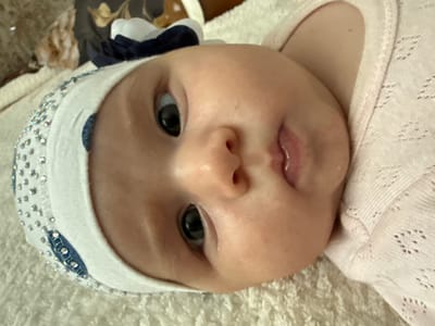 A close-up, overhead shot shows a baby lying on its back on a textured white surface, possibly a blanket or towel. The baby is wearing a white headband adorned with silver rhinestones and a dark blue bow. The baby's face is the central focus, with its left eye wide open and looking towards the camera. The baby is also wearing a light pink, knitted garment. In the background, out of focus, is a dark object with floral patterns. The scene is indoors, with no discernible city or location details from Serebryakovka, Russia, or any other specific geographic area.
