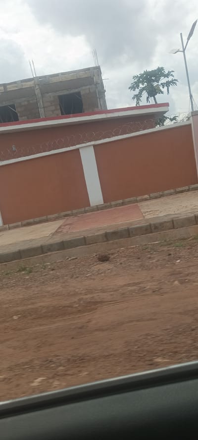 The image shows a two-story building under construction in Umuoye, Nigeria. The building features exposed rebar extending from the roof. In front of the building is a high wall painted brown with white vertical accents and topped with barbed wire. A small tree grows behind the wall, near a street light. The ground is unpaved and reddish-brown. A partially visible car window frame is at the bottom edge of the photo.