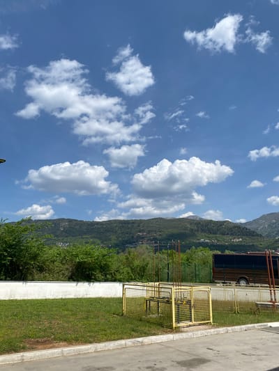 This outdoor image captures a picturesque scene under a bright, sunny sky with scattered white cumulus clouds. The dominant feature in the background is a range of lush, green mountains, heavily forested with trees. Faintly visible on the distant mountain slopes are small buildings, indicating some human settlement.

In the midground, a strip of vibrant green grass runs horizontally, bordered by a white concrete wall or barrier. Behind this barrier and grass, a prominent yellow metal mesh fence or barrier encloses an area. Within this fenced-off section, several tall, brown, pipe-like structures are visible, suggesting an industrial or utility complex, possibly related to gas or water infrastructure. A large, dark brown truck or trailer is partially visible behind the yellow fence.

The foreground on the right features a paved area, likely a road or parking lot, bordered by a concrete curb. No human figures or discernible activity are present in the image. The time of day appears to be bright daylight, given the clear blue sky and strong illumination. There is no visible text. The scene offers a blend of natural beauty with man-made infrastructure, set in Kavač, Montenegro.