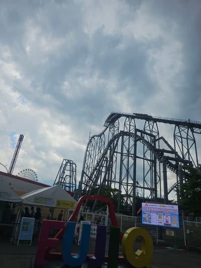 This is an eye-level, medium shot of a roller coaster at an amusement park in Fujikawaguchiko, Japan. The roller coaster dominates the frame, with its intricate metal structure extending into a cloudy sky. A Ferris wheel and a tall, red tower are visible in the distance. In the foreground, a colorful sign spells out "FUJIQ" in large, block letters. White tents and a digital display board with Japanese text are also present. Several people are visible near the tents, some appearing to be staff members and others patrons. The overall atmosphere suggests a theme park environment.