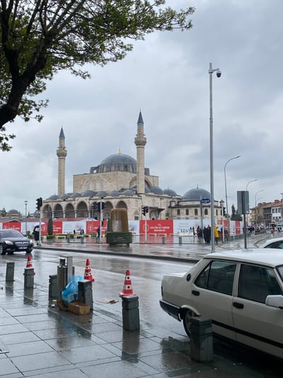 This image presents a view of a wet street in Karatay, Turkey, dominated by the facade of a mosque. The mosque features multiple domes and minarets. Banners labeled "T.C. KÜLTÜR VE TURİZM BAKANLIĞI" are prominent along the street, likely indicating sponsorship by the Ministry of Culture and Tourism. Orange-and-white traffic cones mark the edge of the street along with concrete blocks. A white car is parked on the right edge of the frame, partially blocking the view of the mosque and wet street. A few people are visible in the background. The sky is overcast, suggesting rainy weather. A tree branch is on the left edge of the frame with few leaves. The license plate on the car reads "42 NB 773". A streetlight with a security camera is also in the frame.