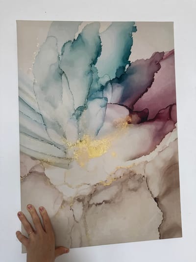 This is a media file showing a rectangular abstract painting held by a person's hand. The painting features flowing watercolor-like patterns in shades of pink, blue, teal, gold, and beige, with a marbled texture. The person's hand, with nail polish and a ring, is placed on the lower right corner of the painting, as if to hold or present it. The background is a plain white surface, likely a wall. There is no visible text in the image. The lighting suggests an indoor setting.
