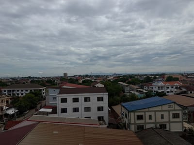 The image is an aerial view of a cityscape in Vientiane Capital, Laos, on an overcast day. The scene is dominated by a dense collection of buildings, predominantly residential and commercial structures with varied architectural styles. Rooftops of corrugated metal and tiles, in shades of brown, red, and grey, are prominent in the foreground and middle ground. Several multi-story buildings, some painted white with dark-framed windows and others in lighter colors, are visible. One building features a bright blue, sloping roof. Trees and vegetation are interspersed throughout the urban landscape, suggesting a tropical climate. In the distance, a hazy horizon shows more buildings and possibly hills or mountains. The sky is filled with a thick layer of grey clouds, obscuring any direct sunlight. No people or vehicles are clearly discernible, giving the impression of a quiet, uneventful moment in the city. The overall impression is one of a densely populated urban area under a cloudy sky.