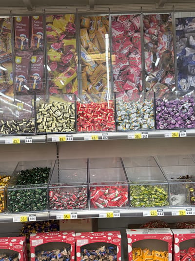 The image shows a retail display of bulk candies in plastic bins at a supermarket in Sokilnyky, Ukraine. The candies are of various colors and wrappers, including red, yellow, purple, and green. Some of the wrappers have text that is difficult to read but appears to be in Ukrainian. Below the bins are price tags with currency and numbers, indicating prices per unit weight. The environment appears to be an indoor market or grocery store with overhead lighting and metal shelving. There are no people visible in the image. The overall scene is one of a commercial setting focused on the sale of confectionery.