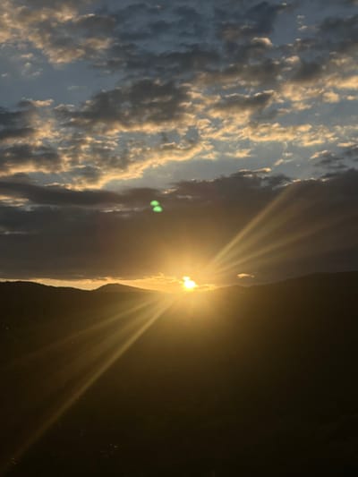 A sideways view captures the sun setting over a dark landscape. Golden sunbeams radiate outward, partially obscured by a jagged horizon line. The sky transitions from dark to a band of white clouds tinged with gold. Additional cloud formations, tinged with silver and gray, fill the left portion of the frame. Two green spots of light are also visible.
