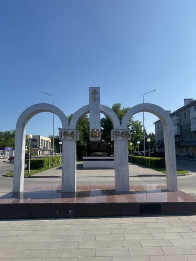 This is a forward-facing, eye-level, full shot of a war memorial in Belovo, Russia. The memorial consists of three large, concrete archways, the central archway is taller than the two side archways. On the central archway, a gold-colored orthodox cross is mounted at the top, and below it, a gold-colored star is visible. The years "1941" and "1945" are displayed in gold lettering on the side pillars of the monument. Behind the central archway, a tank is partially visible, indicating this memorial likely commemorates World War II. The memorial is situated on a paved plaza with a granite base. In the background, trees, streetlights, a multi-story apartment building, and a commercial building with a gas station are visible. The sky is clear and blue. No people are clearly visible in the immediate vicinity of the memorial.