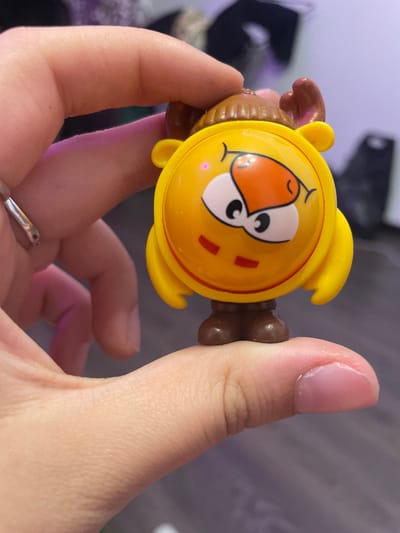 A close-up shot shows a person's thumb and index finger holding a small, yellow, anthropomorphic toy. The toy has a round head with large white eyes featuring black pupils and red accents. It has a large orange nose, and two small red rectangles on its cheeks. The toy wears a brown hat with ribbed texture and two brown reindeer antlers. It has yellow ear-like protrusions on the sides of its head and tiny brown feet. The background is blurred, suggesting an indoor setting with subdued lighting and hints of purple and black. There is no identifiable information indicating the location in Borisova Griva, Russia.