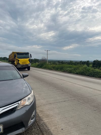 In Oduape, Nigeria, on a cloudy day, a yellow dump truck with visible front-end damage is parked on the right side of a paved road. To the left of the truck, the front portion of a dark gray car is visible, also parked on the side of the road. The road stretches into the distance, bordered by lush green vegetation and trees on both sides. In the background, a utility pole with wires stands against the overcast sky, which is filled with dramatic cloud formations. The overall scene suggests a roadside stop, possibly due to the truck's apparent damage.