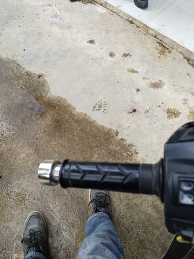 The image shows a first-person perspective of someone standing on a wet, grey concrete surface. The concrete is stained with dirt and there are several small, dark, circular imprints on it, possibly from water droplets or debris. The person is wearing dark-colored sneakers with laces and blue jeans. In the foreground, a black motorcycle handlebar with a textured grip and a chrome end is visible. The handlebar has some controls on the right side, partially obscured. The scene appears to be outdoors, possibly in an urban or semi-urban environment.