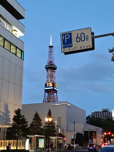 The image depicts a scene in Sapporo, Japan, featuring the Sapporo TV Tower in the background. A modern building, likely an office or commercial structure, is on the left side of the frame, with its windows illuminated. A parking sign with Japanese text indicating "60 minutes" and "Parking Ticket" is visible on the right, mounted on a pole. The Sapporo TV Tower stands tall, displaying the time "7:35" in large, illuminated digits. Below the time display, a "Panasonic" logo is visible. In the foreground, there are trees and a bus stop shelter. Several people are seen near the bus stop and on the sidewalk. A blue car is driving on the street in the lower right portion of the image, with traffic lights also visible. The sky is overcast and the lighting suggests it is dusk or evening.
