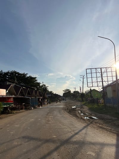 The image depicts a dusty, unpaved road in Alurcucur, Indonesia, lined with small, modest buildings and shops. The sky is a bright blue with scattered clouds and the sun is shining, casting long shadows. Several motorcycles are parked along the side of the road, and a few are visible in motion. Some of the shops appear to be selling goods, with items displayed outside. A person is seen hosing down the road in front of one of the shops. There are also utility poles with electrical wires crisscrossing overhead, and a large, empty billboard structure. Puddles of water and mud are present on the road surface, indicating recent rain or poor drainage. The overall impression is of a busy, yet somewhat dilapidated, rural or semi-urban street scene.