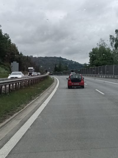 The image depicts a highway scene with a cloudy sky, suggesting overcast weather possibly with rain, as indicated by the water droplets on the windshield. In the foreground, the right lane of a multi-lane highway is visible. A red hatchback car is in the right lane, and its license plate is partially obscured. In the distance, in the left lane, a white car is partially submerged in water, with its headlights on. Further down the highway, a semi-truck and other vehicles are traveling. The highway is flanked by a grassy embankment on the left and sound barriers on the right. The landscape behind the highway consists of wooded hills. The location is identified as the D1 highway near Hvězdonice, Czechia, a known area for water accumulation on the roadway.