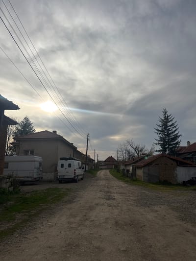 The image shows a dirt road in the village of Gromshin, Bulgaria. The road is lined with buildings, including houses and sheds, and a few evergreen trees are present on the streetscape. A white camper van and a caravan are parked on the side of the road. Electrical wires run above the street. The sky is overcast. The sun is visible and low in the sky, suggesting it is either early morning or late afternoon. No people are visible.