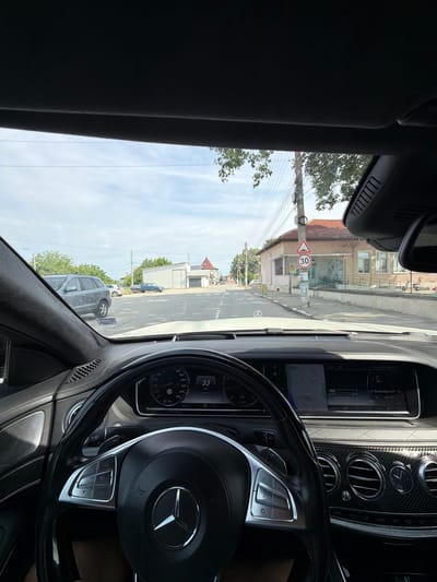 The image captures a first-person perspective from the interior of a Mercedes-Benz car, looking out onto a sunny street scene in Miriantsi, Bulgaria.

Inside the car, the multi-function steering wheel with the prominent Mercedes-Benz logo and silver controls is visible in the foreground. The dashboard's instrument cluster shows a speed of "33 km/h", an engine temperature of "90°C", and a fuel level at "25%". To the right, the car's interior features stylish air vents, carbon fiber trim, and a quilted design on the passenger side dashboard. The infotainment screen displays the time "10:05", along with Cyrillic text "Yattel B Po" and "Излей Духа Си/К", and other details like "01:20", "06:57", and "Mario's iPhone".

Outside the vehicle, a street is visible under a bright, partly cloudy sky. On the left side of the road, buildings with red tiled roofs and white facades line the street. A prominent triangular road sign with a red border and a black number "30" indicates a speed limit of 30 km/h. Utility poles with multiple electrical wires run alongside the road. Further down the street, several cars are parked, including an SUV and a blue pickup truck. Green trees are interspersed among the buildings and vehicles. The scene depicts a typical daytime setting in a town or village.