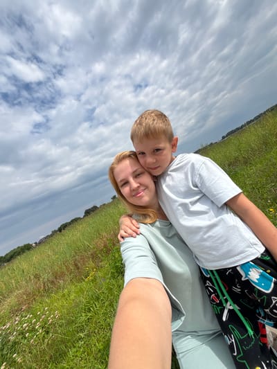 This is a selfie of a young boy and a woman lying on a grassy field. The boy, with blonde hair and blue eyes, is lying on top of the woman, with his head resting on her forehead. He is wearing a white t-shirt and patterned shorts. The woman has blonde hair and is wearing a light blue shirt. They appear to be enjoying themselves, with the woman smiling slightly and the boy looking at the camera.

The background consists of a vast expanse of green grass, with some wildflowers scattered throughout. In the distance, trees and some buildings can be seen, suggesting a rural or park-like setting. The sky is cloudy, with a mix of blue sky and white clouds, indicating it is daytime. The overall mood of the image is cheerful and relaxed, capturing a moment of connection between the two individuals.

The image appears to be taken outdoors in a natural setting, likely during the day. There is no visible text in the image.