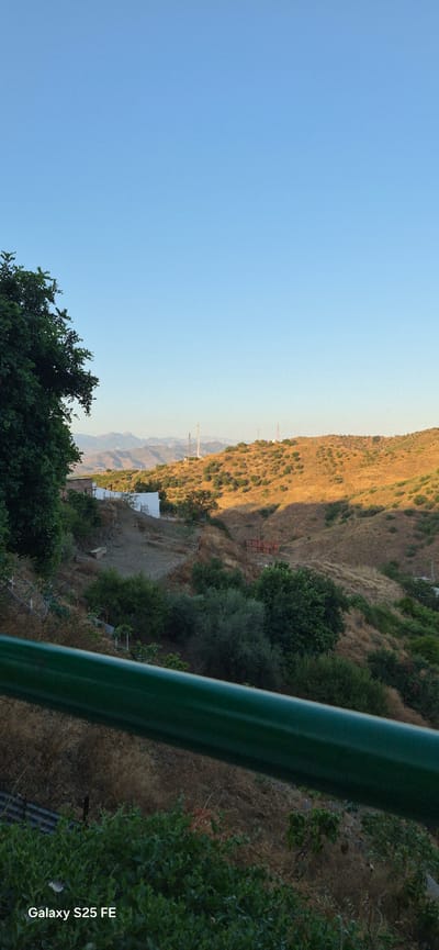 A scenic view of the hills surrounding Almáchar, Spain, is captured under a clear blue sky. In the foreground, a thick, dark green railing runs horizontally across the bottom half of the image. Beyond the railing, the landscape slopes downwards into a valley dotted with various trees and dry, golden vegetation. A small, white building with a dark roof is visible on the left side of the valley, near a fenced area. Further into the distance, the hills are covered in sparse vegetation, with several communication towers standing on the ridgelines. The mountains in the far background are hazy and appear to be layered. The image also contains a watermark in the bottom left corner that reads "Galaxy S25 FE."