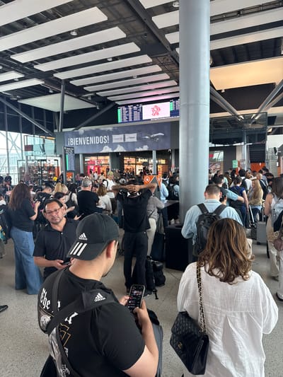 This media file captures a bustling scene inside a modern airport terminal, likely in Apodaca, Mexico, as suggested by the location context and visible cues. The perspective is from behind several individuals in the foreground, looking towards a crowded waiting or check-in area.
Numerous people of various ages are gathered, predominantly standing in lines or congregating. Many are occupied with their mobile phones, either holding them or looking at their screens. Some individuals are seated, while others are standing with luggage, including rolling suitcases and backpacks.
The setting features high ceilings with prominent white rectangular acoustic panels and a grey cylindrical pillar dominating the foreground. Large digital screens display information, some showing what appears to be flight schedules or advertisements. One screen prominently features a red lion-like logo, possibly for an airline like Aeromexico.
Visible text and branding include:
* A large sign proudly displaying "¡BIENVENIDOS!" (Spanish for "WELCOME!").
* On an information screen, "VINCI" and "OMA" (Grupo Aeroportuario del Centro Norte, an airport operator) are visible.
* Another sign indicates gate numbers "22-25".
* The "Adidas" logo is clearly seen on a cap and shirt worn by individuals in the foreground.
* A small sign for "Continental" is also present.
The activity primarily involves waiting, typical for an airport environment. The lighting is bright and artificial, suggesting an indoor setting and no specific time of day like dawn or dusk, but rather general operational hours. The overall atmosphere appears orderly despite the crowd, with people patiently engaged in their personal activities while awaiting their next step in travel.