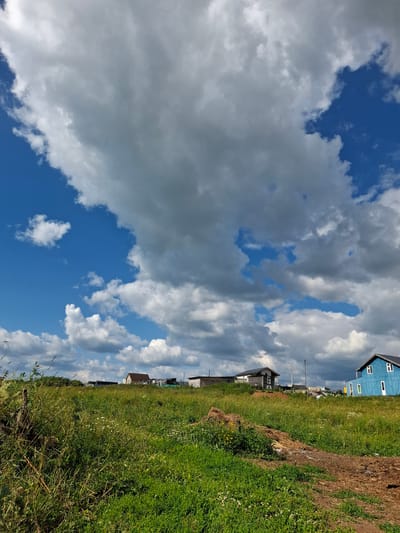 This is a landscape photograph taken on a sunny day with a bright blue sky dotted with fluffy white and gray clouds. In the foreground, a grassy field stretches out, with tall grasses and scattered wildflowers. A dirt path is visible in the lower right quadrant. In the middle ground, there are several buildings, including a brown-roofed house, a darker-colored house, and a bright blue house with white trim and windows. The scene is set in the village of Kosyakovo, Russia.