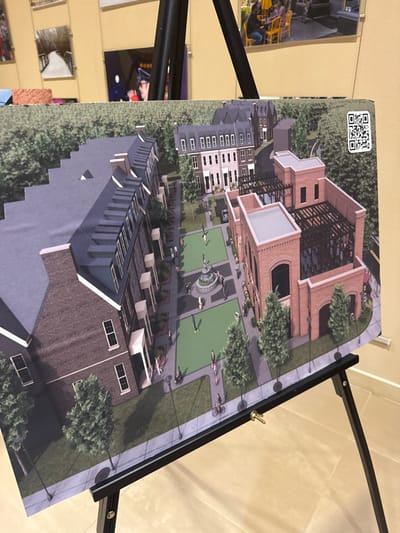 An architectural rendering depicts a planned community. A central green space features a fountain, surrounded by pedestrian paths and people. Townhouses with pitched roofs anchor the bottom of the image. A taller building with a white facade is to the left. A brick building with a modern, open-air top sits at the upper right. A QR code is visible in the upper left corner. The rendering is mounted on a black easel.