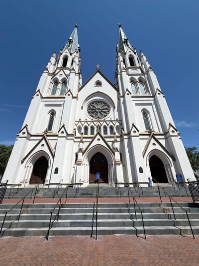 The image shows the exterior of a large, white Gothic-style cathedral with two tall spires and a rose window. The building has intricate architectural details, including pointed arches, finials, and decorative stonework. A wide set of stone stairs leads up to the main entrance, with metal handrails on either side. The stairs are bordered by brickwork. Blue signs with white text are visible on the building. The sky is clear and blue, indicating a sunny day. The sun casts shadows on the building and stairs, suggesting it is daytime. There are no people visible in the image. The scene is set in front of a grand cathedral, likely in Savannah, United States of America, given the location context.