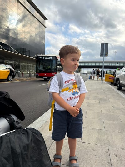 A young boy with brown hair, wearing a white t-shirt with a cartoon image and text, dark blue shorts, and grey sandals, stands on a sidewalk next to luggage. He has a grey backpack with yellow straps. In the background, a red bus is driving on a street, a yellow taxi is parked on the side, and a modern building with reflective windows is visible. The sky is cloudy. The scene appears to be an outdoor area near an airport or transportation hub, potentially in El Prat de Llobregat, Spain, given the context.