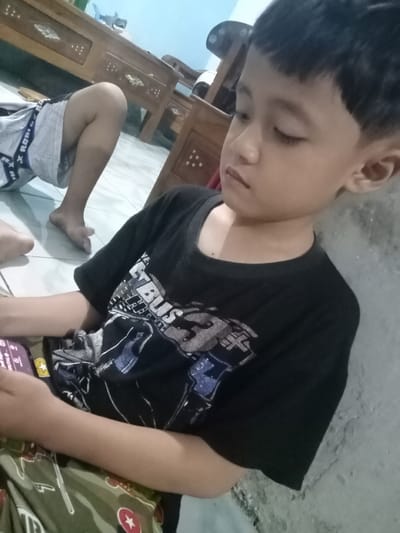 A child with dark hair and a black t-shirt sits indoors, appearing to be looking down, perhaps at a phone in their lap. Another person's leg is visible in the background. The location is likely Bungur, Indonesia. A wooden table is behind them.
