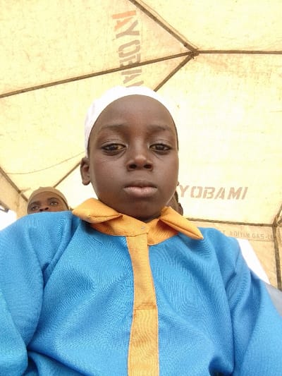 A frontal, close-up image captures a child, appearing to be of primary school age, with dark skin, wearing a white kufi cap and a blue long-sleeved garment with an orange pointed collar and a central orange strip. The child looks directly forward with a neutral expression. Partial views of at least two other individuals are visible behind the primary subject; one to the left and rear wears a light brown head covering, with only the forehead and eyes discernible. The background is formed by a light-colored, possibly beige or yellow, fabric canopy, indicative of a tent or temporary shelter. Inverted and mirrored text is visible on the canopy, including "IYORI," "YOBAMI," and "ADIYAN GAS-L," which appear to be branding or signage. The presence of "ADIYAN GAS" suggests a local business, consistent with a setting in Orudu-Olokan, Nigeria. The scene is illuminated by diffused light from above.