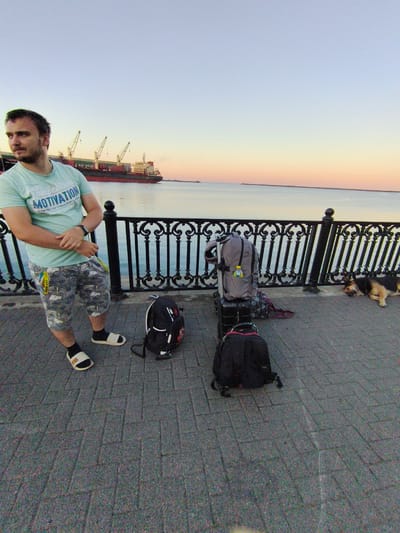 A man stands on a paved walkway next to a railing overlooking a body of water. He is wearing a light blue t-shirt and camouflage shorts, with white slides on his feet. To his right are two backpacks, one black and one gray, and a suitcase. A dog sleeps on the walkway near the railing. In the background, a large cargo ship is docked in the water, with cranes on its deck. The sky is a gradient of blue and orange, indicating either sunrise or sunset. The scene is consistent with a waterfront promenade in Agoy, Russia, a coastal town known for its port and recreational areas.