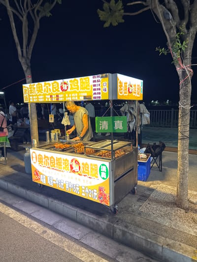 The image shows a food stall at night in Eshan Yi Autonomous County, China. The stall is selling chicken legs, as indicated by the Chinese characters on the signage which translate to "New Erliang Chicken Legs." The stall has a grill with chicken legs cooking on it, and a man, presumably the vendor, is preparing food. There are customers in the background, suggesting a busy street food scene. The environment appears to be a paved street with trees and a body of water in the distance. The lighting suggests it is after dark, with the food stall and its signage being the primary light sources.