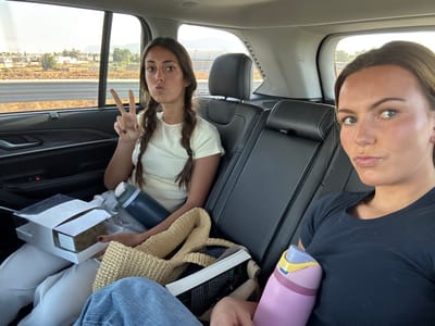 The scene depicts two young adult females seated in the back of a vehicle, likely traveling or parked in Tultepec, Mexico, during daylight hours. The female on the left, with braided brown hair, wears a light-colored short-sleeved top and light-colored pants. She has her left hand raised, displaying a two-finger "peace" sign, and her lips are pursed. On her lap are a dark-colored insulated water bottle, an open white cardboard box with a transparent lid containing a food item, and a light-colored woven bag. The female on the right, with brown hair, wears a dark-colored short-sleeved top and blue jeans. Her lips are also pursed, and she holds a pink and blue insulated water bottle against her chest. Both individuals are looking towards the camera. The vehicle's interior features dark leather seats and dark door panels. Through the windows, a dry, open landscape with scattered low-rise buildings, distant hills, and a road barrier is visible under a clear sky.