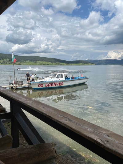 This image depicts a daytime scene at the Цигов чарк reservoir in Bulgaria, under a partly cloudy sky with prominent cumulus clouds and patches of blue. The water surface shows slight ripples and some floating debris or foam in the foreground.
In the midground, a white and blue motorboat is docked at a wooden floating pier. The side of the boat bears the Cyrillic inscription "ОХ БАБАЧКО" in red capital letters, with its reflection visible on the water. A Bulgarian flag, horizontally striped white, green, and red, is affixed to a pole on the dock near the boat's stern. Three individuals are visible on the dock or boat platform: one man, seen from behind wearing a black shirt and dark shorts, stands near the edge of the dock, and two younger individuals, in light-colored shirts and shorts, are also present on the dock.
Further out on the water to the left, a jet ski is actively moving, generating a white wake. The background consists of rolling hills densely covered with green forests, extending to the horizon under the cloud-filled sky. The photograph is taken from an elevated perspective, looking over a wooden railing that spans the lower portion of the image, suggesting the vantage point is from a lakeside structure or terrace.