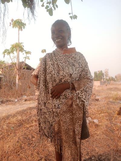 A young individual with dark skin and braided hair stands smiling outdoors in Gudige Sabo, Nigeria. They are wearing a poncho-style garment featuring a leopard print and a central reptilian-scale pattern, with a fringed hem and an exposed orange-brown collar. A segmented metallic bracelet is visible on the left wrist. The right hand rests on the abdomen. The dry, reddish-brown ground is sparsely covered with dead vegetation and minor debris. In the background, a tall, slender tree with minimal foliage stands left of a dilapidated building. The sky is hazy. Overhead foliage and thin roots are visible in the upper left corner of the frame.