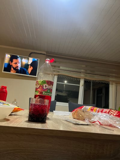 A bottle of raspberry drink (Derby brand) and a glass filled with red liquid are on a wooden table. A television screen shows a man with a beard. The image was likely taken indoors, near a window covered with curtains. The location of Slivo Pole, Bulgaria cannot be verified from this image.