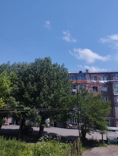 The image depicts an exterior view of a multi-story apartment building in Hrazdan, Armenia. The building, constructed with reddish-brown stone, features multiple windows and balconies, some of which have laundry hanging from them. Two large, leafy trees dominate the foreground, their branches partially obscuring the building. The sky above is a clear blue with a few scattered white clouds. A power line stretches across the upper portion of the image, and a metal fence is visible in the lower right corner. The ground in front of the building appears to be a paved area, possibly a courtyard or street, with some patches of green vegetation and white wildflowers. A dark-colored vehicle is partially visible under the shade of the trees. The scene conveys a typical residential urban environment on a sunny day.