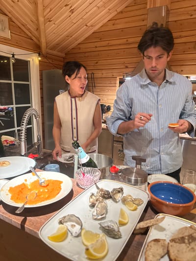 This image captures an indoor scene in a kitchen located in Boda, Sweden. The environment features walls and an angled ceiling constructed from light-colored wooden planks, consistent with a log cabin or rustic home interior.

Two individuals are present: an adult female on the left and an adult male on the right. The female has dark hair pulled back and wears a cream-colored sleeveless top with dark trim and gold buttons; her mouth is slightly open, and she is looking towards the male. She holds a small white plate with a single oyster. The male has dark, wavy hair and wears a light blue striped long-sleeved button-up shirt; he is looking down intently at a small round blini or cracker topped with orange roe, which he holds in his left hand. A ring is visible on his left hand.

A variety of food items and servingware are arranged on a wooden countertop in the foreground and midground. These include a large white plate containing a substantial amount of orange roe or caviar with a silver spoon, a separate white plate with a single pancake or blini, and a rectangular white platter displaying multiple raw oysters on the half-shell garnished with lemon wedges. A metallic champagne bucket holds a light blue-labeled champagne bottle. Adjacent to it is a small, clear glass bowl containing red finely chopped contents, possibly onion. Other items on the counter include a light blue beverage glass, a brown and blue ceramic bowl, a metal shaker/grinder, an empty champagne flute, and a white plate with slices of dark bread.

In the background, a stainless steel refrigerator is partially visible to the left, and a modern kitchen sink with a spring-coil faucet is also present. A stove or oven unit is partially visible to the right. String lights are strung above a window on the far left.