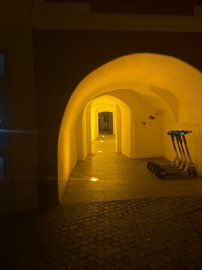 The image displays an illuminated, arched stone passageway at night within the Old Town of Sibiu, Romania. The immediate foreground shows a dark, irregular cobblestone surface. The passage itself comprises a succession of light-colored, stuccoed arches, creating a tunnel effect that recedes into the distance. The interior of the passage is brightly lit by warm, yellow-orange light from multiple circular, recessed ground lights embedded in the smoother, rectangular paving stones that transition from the cobblestone entrance. Against the right wall, approximately midway into the brightly lit section of the passage, four white electric scooters are parked upright, displaying glowing blue lights on their handlebars. The right wall also exhibits some minor circular marks or superficial damage. A partially visible "glo" sign is present on the dark wall segment to the immediate left of the passage entrance. Further down the passage, a "P" (parking) sign is partially discernible on the left side. No individuals are present in the scene, and no explicit actions or interactions are depicted. The environment is urban and characteristic of a historic pedestrian thoroughfare.