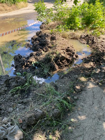 The image depicts a disturbed area with muddy water and piles of soil, suggestive of recent excavation or flooding. A warning tape with red and white stripes is visible across a muddy puddle, indicating a potential hazard or restricted area. Lush green vegetation surrounds the scene, and a dirt road is partially visible in the background. The overall impression is one of disarray and possible ongoing repair or construction work in a semi-rural or unpaved urban environment.