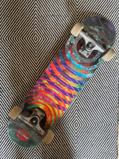 A skateboard with a colorful, cosmic-themed design lies diagonally on a black and white striped rug with a diamond pattern. The skateboard has silver trucks and tan wheels. The rug is textured with thick woven threads. The scene appears to be indoors, likely on a floor. The image is taken from a top-down perspective.