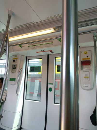 The image depicts the interior of a modern Lima Metro Line 1 train car at Villa El Salvador station. The car features white walls and ceiling, illuminated by linear LED light strips, some warm yellow and others cool white. AC vents are visible on the ceiling. Brushed metal vertical poles and horizontal handrails provide passenger handholds.

Centrally located double doors are partially open, white with black-framed glass panels. To the left, a panel displays an "Aló LÍNEA 1" sticker (0800-111-21) and an emergency handle labeled "Desbloqueo de puertas / Doors unblocking." Between the doors, a green button reads "Para abrir las puertas / To open the doors." To the right, a control panel features a yellow "Alarma Alarm" label above a red pull handle, with an "SOS" bell icon above it. Below this, an "Ayuda / Help" panel includes a speaker, a button, and instructions, notably "Pulsar para hablar. Fixed light. Press to talk." Above the doors, a red-bordered sign reads "¡Atención! Attention!" warning against entering/exiting when door lights flash.

Above the doors, a strip map for "línea 1 Metro de Lima" displays stations, with "Villa El Salvador" highlighted in red.

On the far left, a person with dark hair and a light, patterned shirt is partially visible behind a pole. An indistinct form is to the right.

Through the open doors, a metro station platform is visible under bright daylight, with "EL SALVADOR" clearly displayed in large red capital letters on the platform wall.
