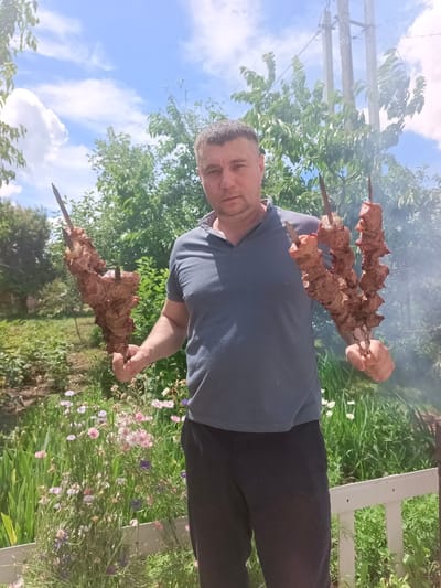 A man is standing outdoors in what appears to be a yard or garden. He is holding two skewers of meat, which appear to be beef, in front of him, one in each hand. The meat is raw and has been cut into chunks. The man is wearing a blue polo shirt and black sweatpants. He has short brown hair and is looking directly at the camera. Behind him, there are green trees and a blue sky with white clouds. There is also a white fence in the foreground. The image is likely taken in Vyazovka, Russia, as indicated by the user's prompt, though this cannot be definitively determined from the image itself. The overall scene suggests a casual outdoor gathering or a barbecue.