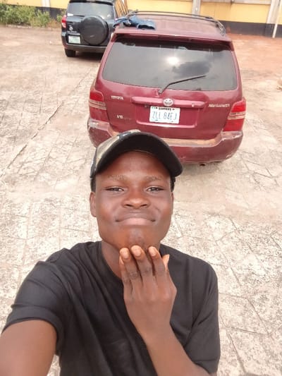 A young man, appearing to be of African descent, is depicted in a selfie-style photograph taken outdoors in Itu, Nigeria. He wears a black t-shirt and a camouflage-patterned baseball cap, worn backward. His left hand is positioned with fingers together under his chin. He exhibits a subtle smile. Behind him, two sport utility vehicles are parked on a paved surface composed of interlocking light-colored concrete blocks. The primary vehicle directly behind the man is a dark red Toyota Highlander V6 with Nigerian license plate "CROSS RIVER ZLL 84EJ." The vehicle displays visible scuffs on its rear bumper and a blue fabric item draped over its roof rack. Further in the background, behind the Highlander, a dark-colored Toyota RAV4 is partially visible, featuring a spare tire mounted on its rear door and a partially obscured license plate. A light-colored building wall is discernible in the distant background. The scene is illuminated by natural light.