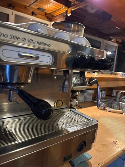 A close-up, upward-angled shot focuses on the control panel and various components of a "Uno Stile di Vita Italiano" espresso machine. The machine, primarily composed of stainless steel, is equipped with black knobs and levers. Various chrome parts and accessories, including a steam wand, are visible in the background. The machine sits on a wooden surface. In the periphery, parts of a building with white-framed windows are faintly visible. The location is unidentifiable as Vitez, Bosnia and Herzegovina, based on this image alone.