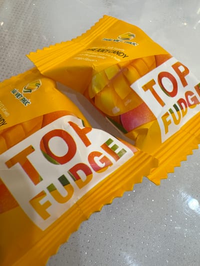 The image shows two yellow candy wrappers with the words "TOP FUDGE" and a mango image. The wrappers are laying on a speckled gray surface, possibly a countertop. There are no people, animals, or indications of the city of Chuguyevka, Russia, visible in the image.