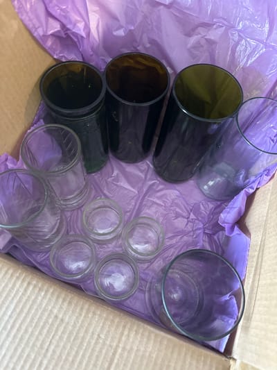 The image displays an assortment of glass containers packed inside a cardboard box, which is lined with crinkled purple tissue paper. The scene is likely indoors in Kuda, Russia.
The objects in the box consist of various types of glass items. On the left side, there are five dark, possibly brown or dark green, cylindrical glass containers, seemingly repurposed from bottle bottoms, lying horizontally. These are uniform in size and shape, suggesting they might be used as glasses or small vases.
To the right, there are numerous clear glass items. Six smaller clear glass jars are arranged in two rows of three, likely repurposed from food jars. Above these, towards the top right, a larger clear glass tumbler or jar stands upright. Another larger clear glass container is partially visible in the upper central part of the box.
The arrangement suggests the items are either being carefully packed for storage or transport, or have just been unpacked. The crinkled purple tissue paper serves as cushioning, indicating care for the fragile contents. No people are visible, and there are no specific cues regarding the time of day or weather. No discernible text is visible on any of the objects or the packaging. The image captures a still life of repurposed glass items, possibly for craft, home decor, or retail purposes.
