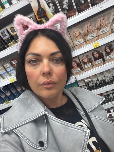 A selfie was taken by a woman with pink cat ears, wearing a silver jacket and black shirt, at a store in Murmansk, Russia. Shelves filled with boxes of hair dye are visible in the background. The hair dye is mostly blonde colors.