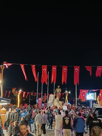 The image shows a gathering of people in Silivri, Türkiye, at the coordinates 41.072, 28.244. The crowd is gathered around a monument of a man in a suit standing on a platform, and the area is decorated with Turkish flags. People are interacting with each other and looking at the monument. There are some food stalls on the left side of the frame. There are also police officers standing in the crowd, wearing uniforms and identification. The event appears to be a public celebration, potentially a commemorative event. The atmosphere is lively and celebratory. The background features a dark night sky with street lights illuminating the scene.