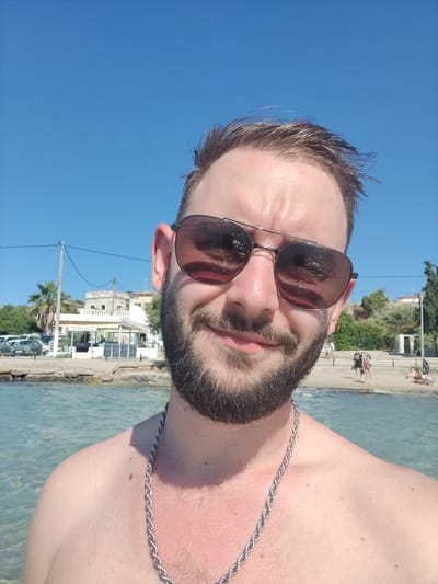 A man with a beard and sunglasses is taking a selfie outdoors. He is shirtless and wearing a thick silver chain around his neck. The man is standing in the water, and the background shows a beach with people in the distance. There are also buildings and palm trees visible. The sky is clear and blue. The image appears to have been taken in Aegina, Greece, judging by the architecture and vegetation in the background. The man is smiling and looking directly at the camera. The water is clear and shallow.