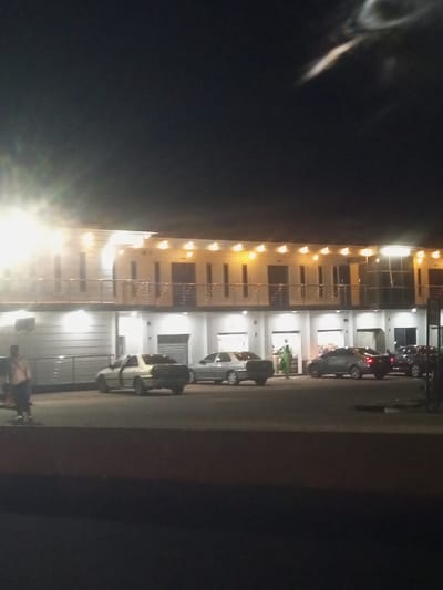 The image depicts a nighttime scene outside a two-story building, likely a hotel or guesthouse, in Baccure Kofare, Nigeria. The building is illuminated by multiple overhead lights, casting a warm glow on its facade. The ground floor appears to have several entrances or open spaces, some with visible interiors. A balcony with a railing runs along the second story.

Several cars are parked in front of the building, including a light-colored sedan with its doors open, a darker sedan with its trunk open, and other sedans. A person wearing a light-colored shirt and dark pants is standing on the left side of the image, appearing to be walking away from the building. Another person, wearing a green garment, is standing near one of the cars.

The lighting suggests it is nighttime. The scene is an exterior view of a commercial establishment, possibly a lodging or a business with parking facilities. There is no visible text in the image. The overall impression is one of a quiet evening outside a building with some activity.