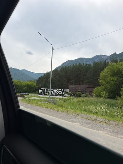 An outdoor scene captures a roadside view with a large hashtag "#TERRASSA" sign made of white block letters against a dark background. To the right of the sign, a two-story brown building with "cafe" written on it is visible, surrounded by trees and a grassy area. A silver car is parked in front of the building. In the background, rolling green hills and a forest of pine trees are seen under a cloudy sky. The image appears to be taken from inside a vehicle, with the dark interior of the car frame visible in the foreground. The location is the city of Izvestkovyi, Russia.